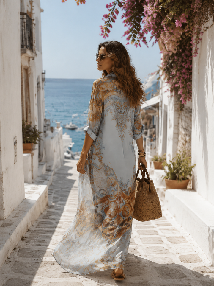 Back view of elegant floral maxi dress in blue tones, worn with a woven bag, overlooking a coastal village and ocean.