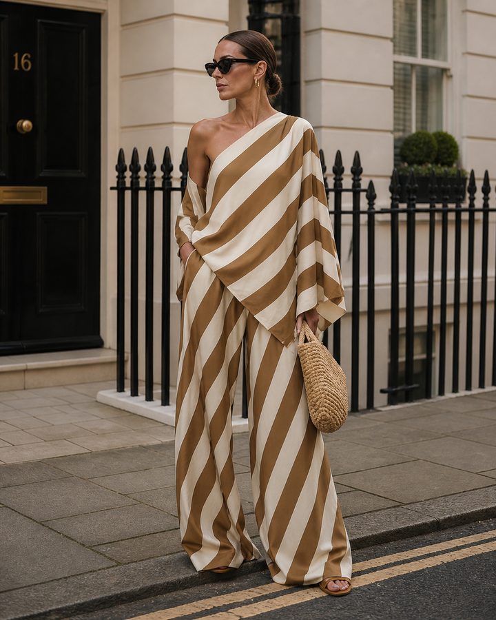 Brown and white striped one-shoulder set worn by model with sunglasses, holding a woven bag, standing on a city street.