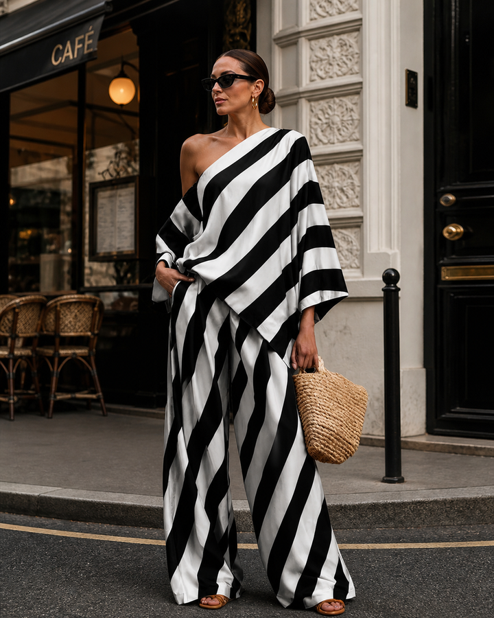 Black and white striped one-shoulder set worn by model with sunglasses, holding a woven bag, standing outside a café.