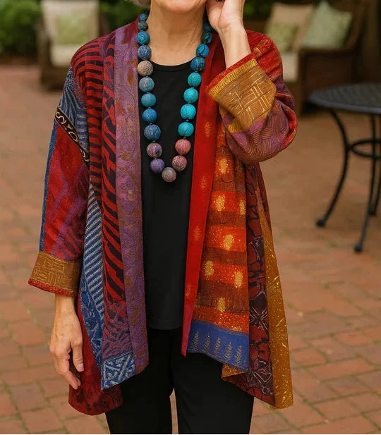 Woman in a richly colored red and blue drape cardigan with intricate designs, posing on a brick patio.