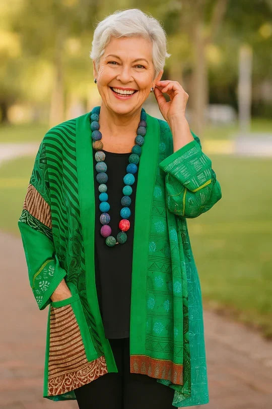 Smiling woman in a green patterned drape cardigan and beaded necklace, posing on a sunny path.