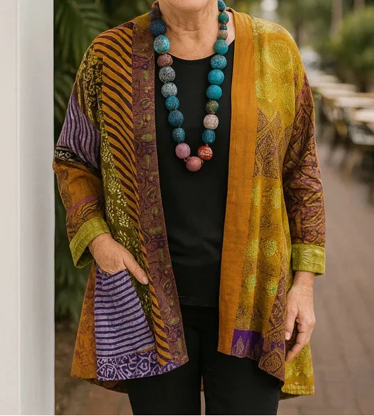 Woman wearing a yellow and purple drape cardigan with vibrant patterns, standing next to a white column outdoors.