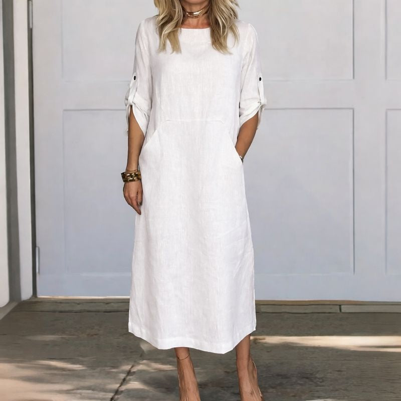 White A-line loose fit draped evening dress, studio setting with wooden floor and pale wall background.