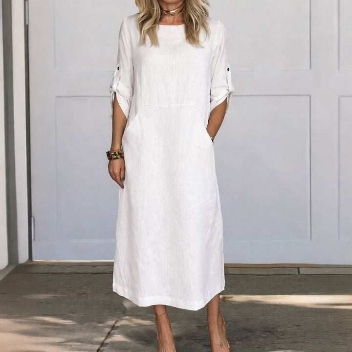 White A-line loose fit draped evening dress, studio setting with wooden floor and pale wall background.