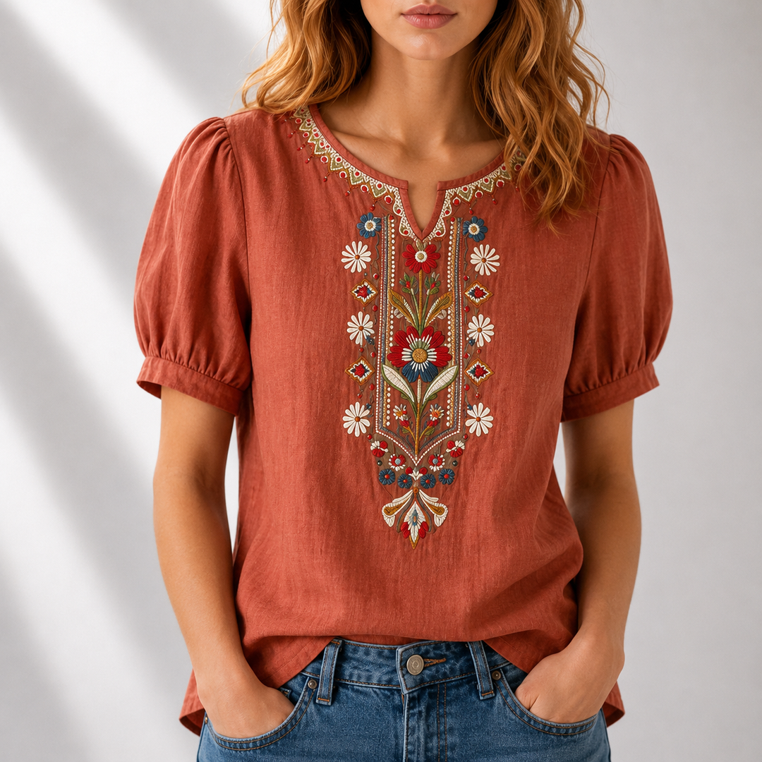 Woman in a rust-colored blouse with floral embroidery, puff sleeves, in a light-filled studio setting.
