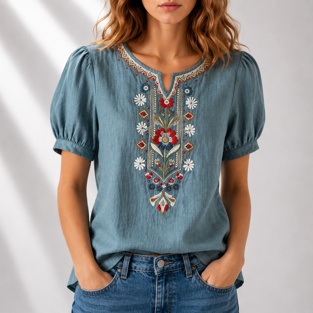 Woman wearing a blue blouse with vibrant floral embroidery, puff sleeves, in a bright studio background.