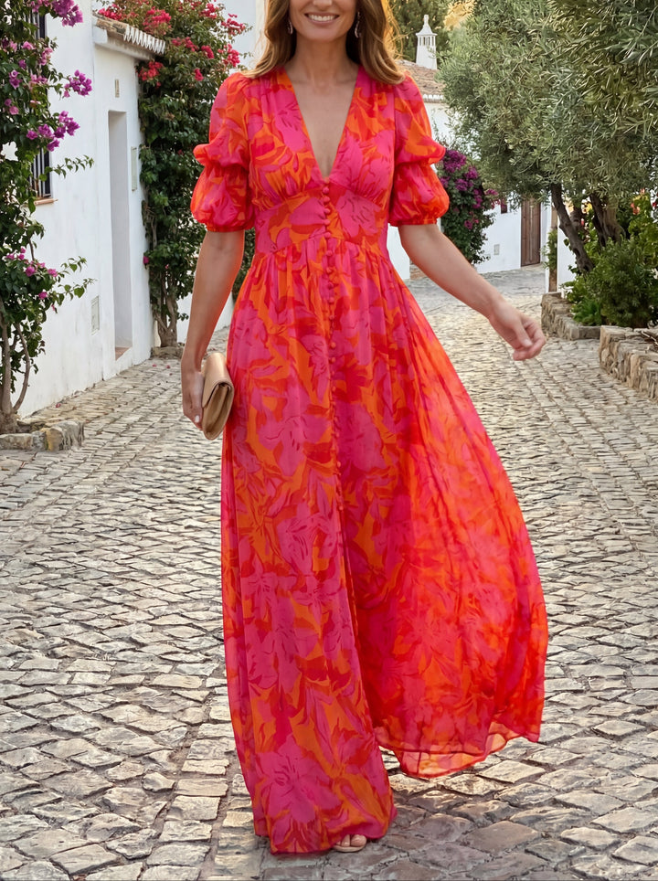Woman in pink and orange floral dress smiling and walking along a quaint cobblestone street with flowering vines.