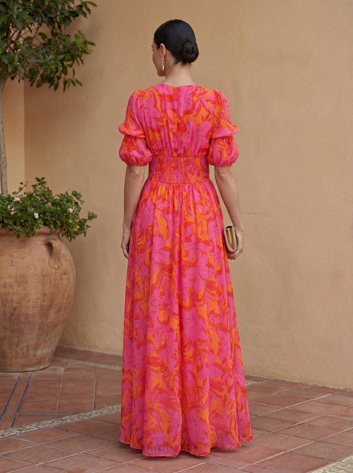 Back view of woman in pink and orange floral dress in courtyard setting with terracotta tiles and a large potted plant.