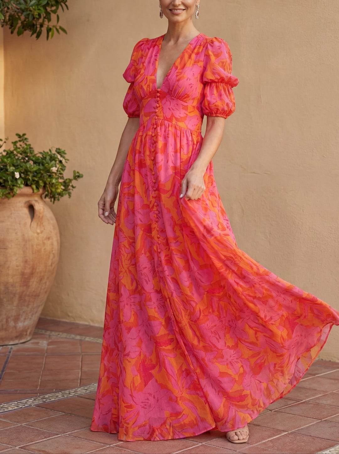 Woman in flowy pink and orange floral dress posing in a tiled courtyard with greenery and terracotta pot.