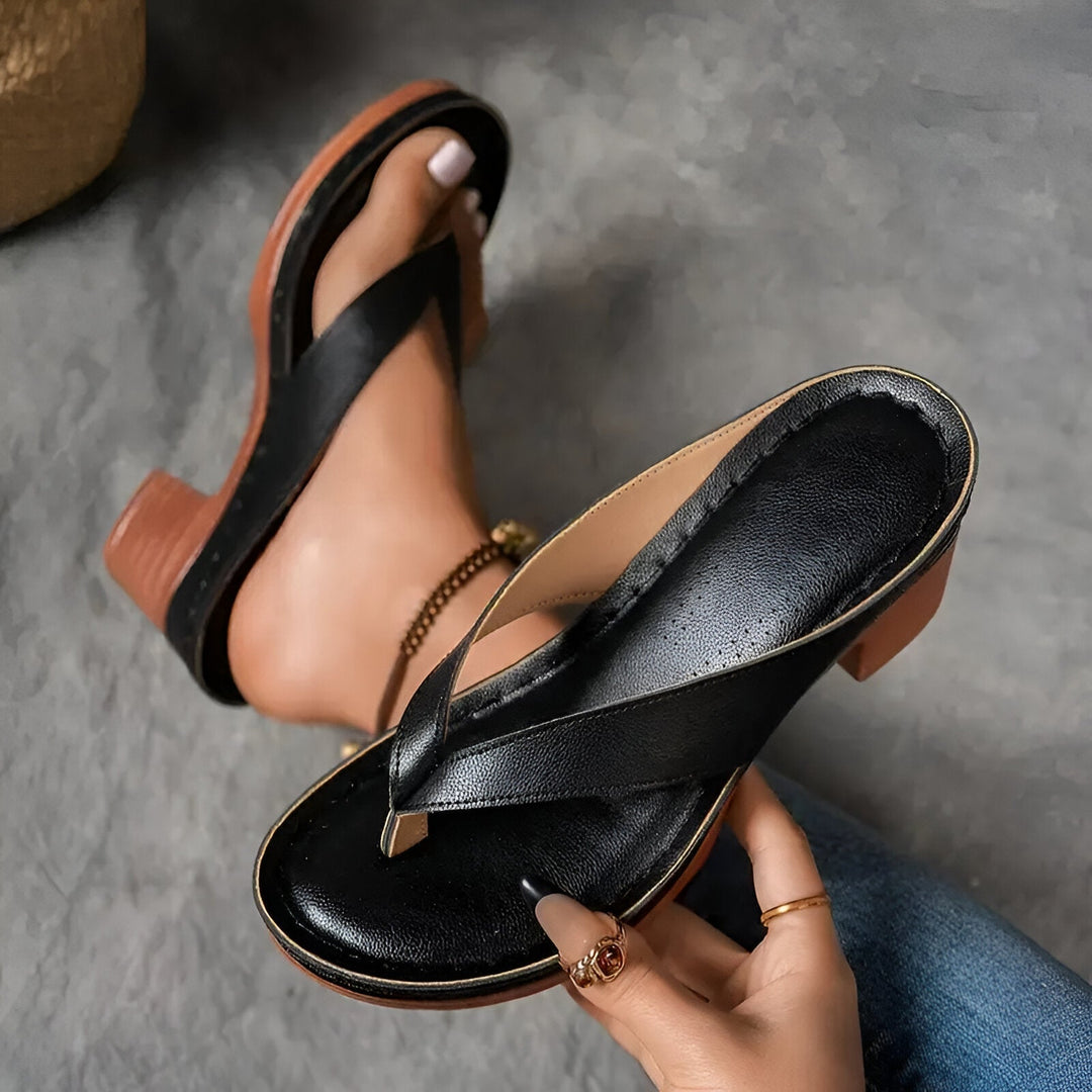 Black high-heeled mule with perforated insole held by a hand, worn on foot with anklet, on gray surface.