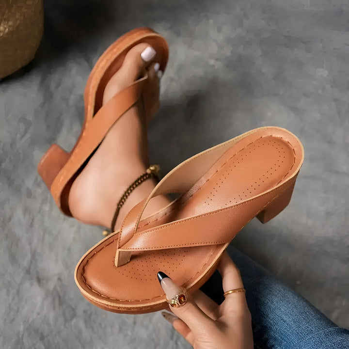 Brown high-heeled mule with perforated insole held by a hand, worn on foot with anklet, on gray carpet.