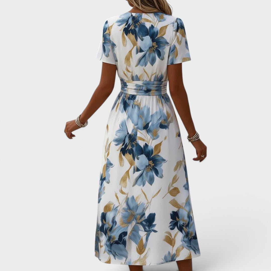 Back view of a woman wearing a floral, blue and gold midi dress with a cinched waist in a studio setting.