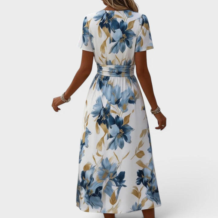 Back view of a woman wearing a floral, blue and gold midi dress with a cinched waist in a studio setting.