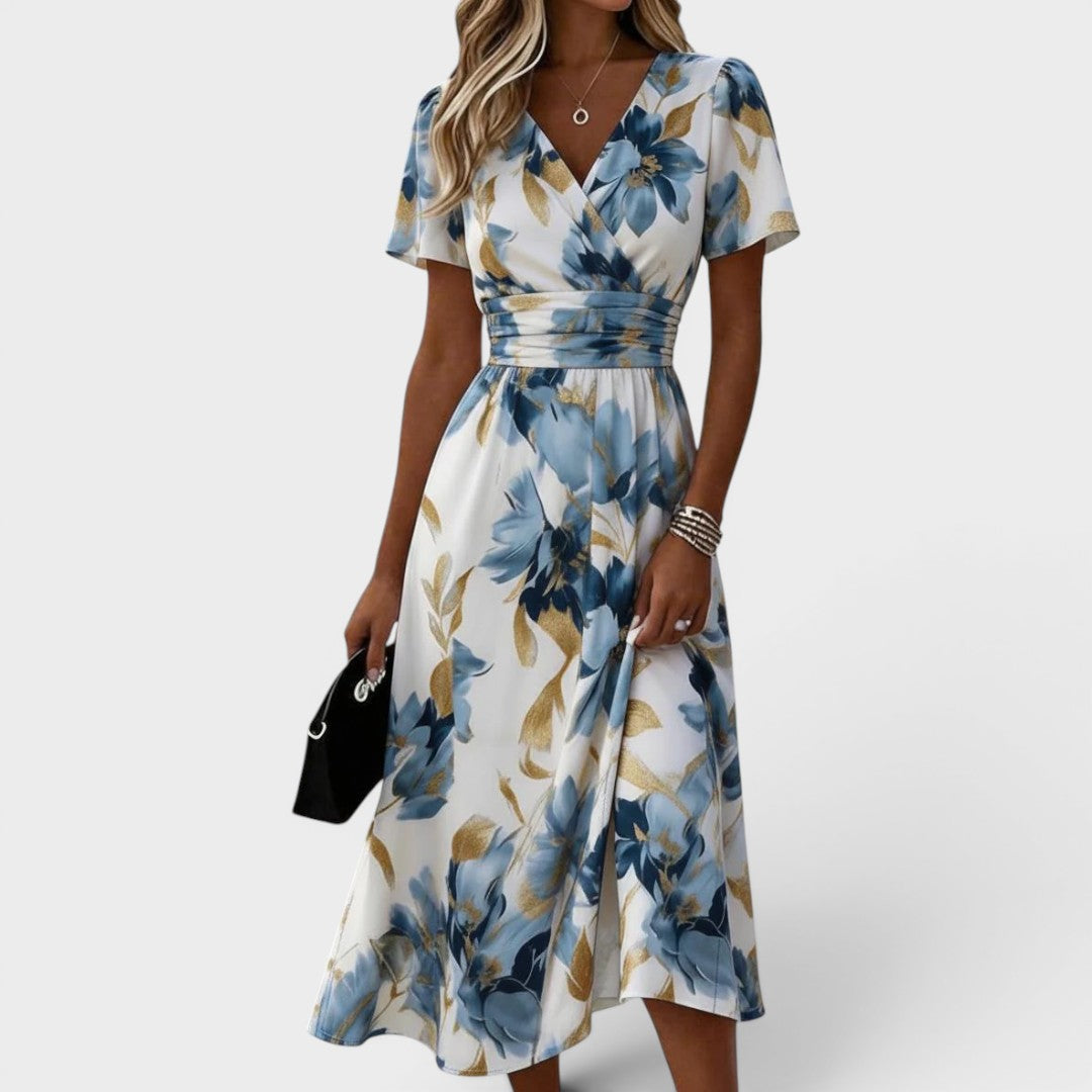 Woman wearing a floral, blue and gold midi dress with short sleeves and V-neck, holding a black handbag in a studio setting.
