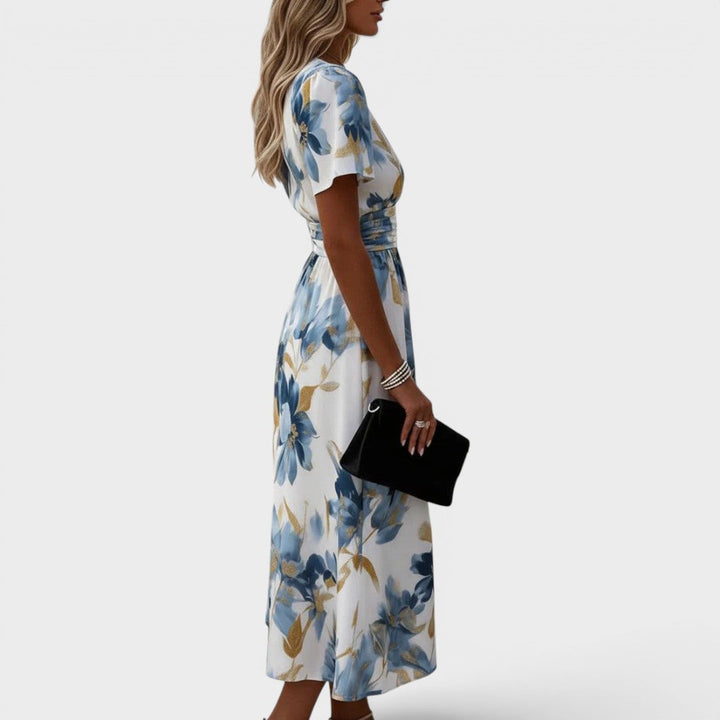 Side view of a woman wearing a floral, blue and gold midi dress, holding a black clutch in a studio setting.