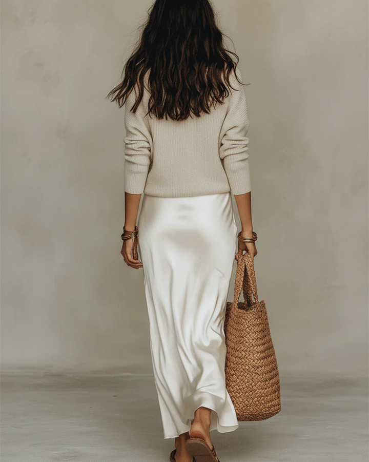 Back view of a woman in a cream sweater and white flowing skirt holding a woven bag, standing on plain background.