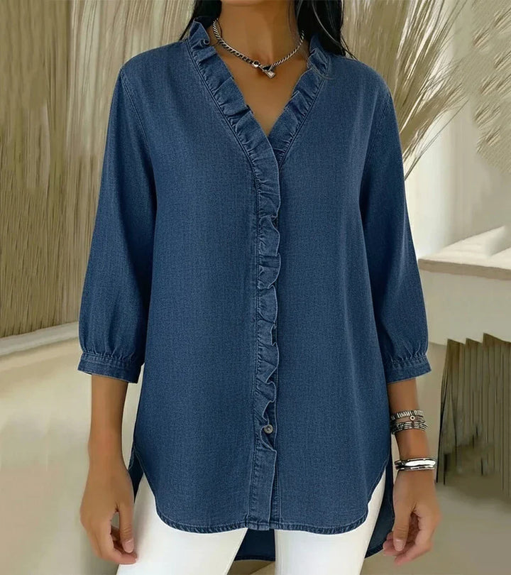 Elegant denim blouse with ruffled trim in blue, worn by model indoors against a neutral background.