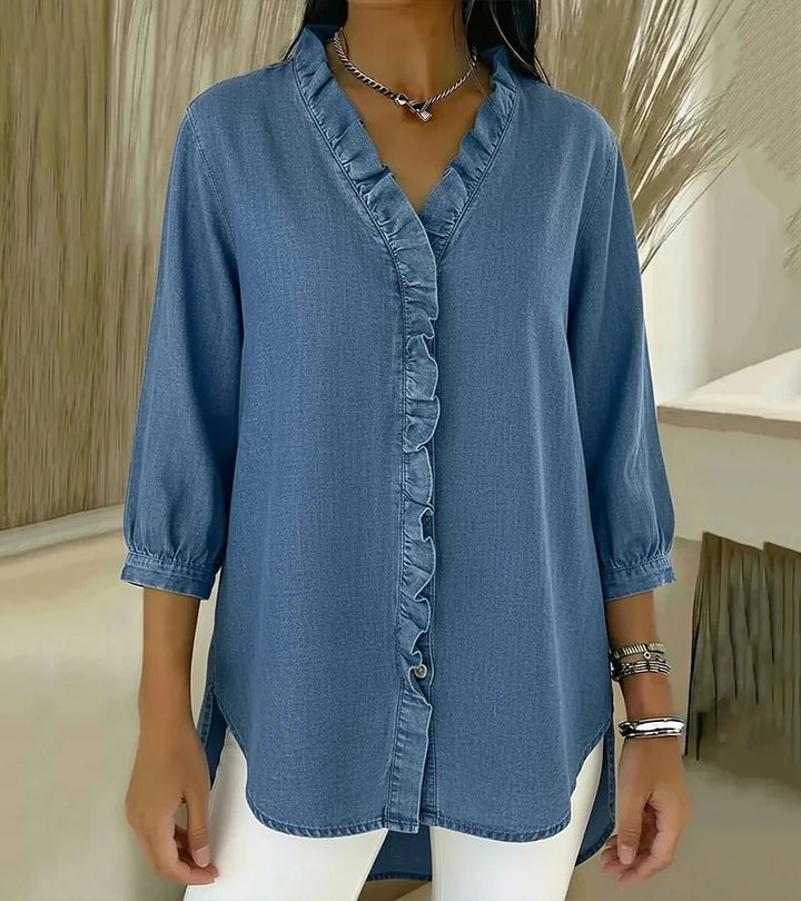 Light blue denim blouse featuring ruffled trim, worn by model indoors with a neutral backdrop.