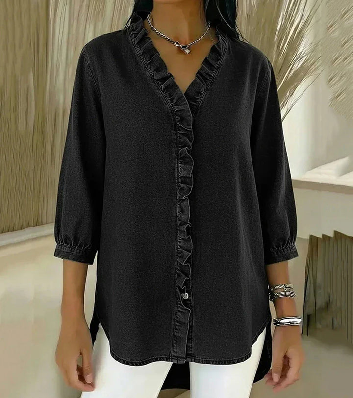 Black denim blouse with ruffled trim and 3/4 sleeves, paired with white pants, indoor setting.