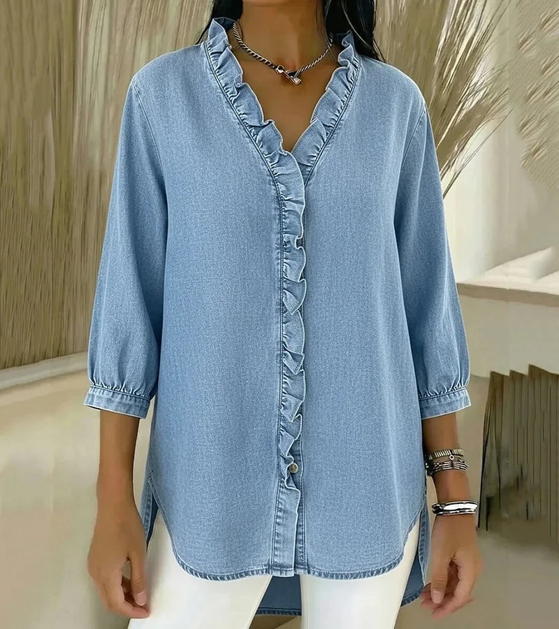 Light wash denim blouse with ruffled trim, styled casually with white pants, displayed indoors.