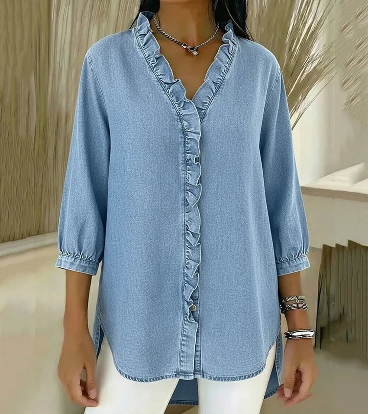 Light wash denim blouse with ruffled trim, styled casually with white pants, displayed indoors.