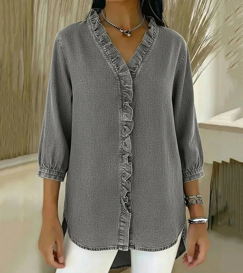 Gray denim blouse featuring ruffled detail down the front, paired with white pants in an indoor setting.
