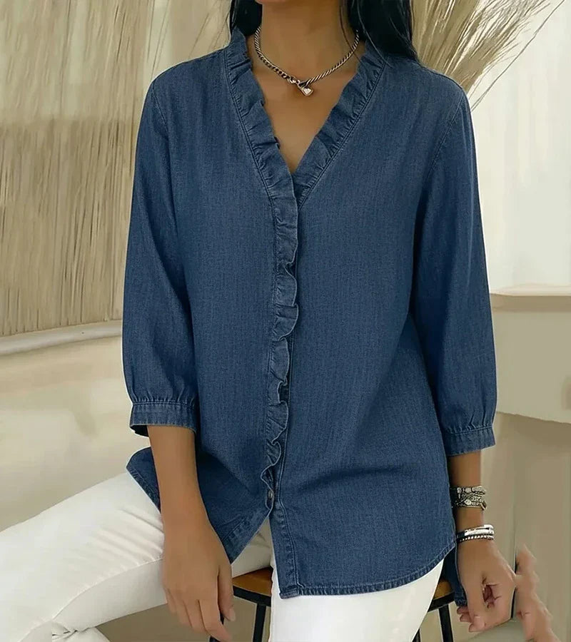 Blue denim blouse with ruffled trim and 3/4 sleeves, styled casually with white pants, indoors.