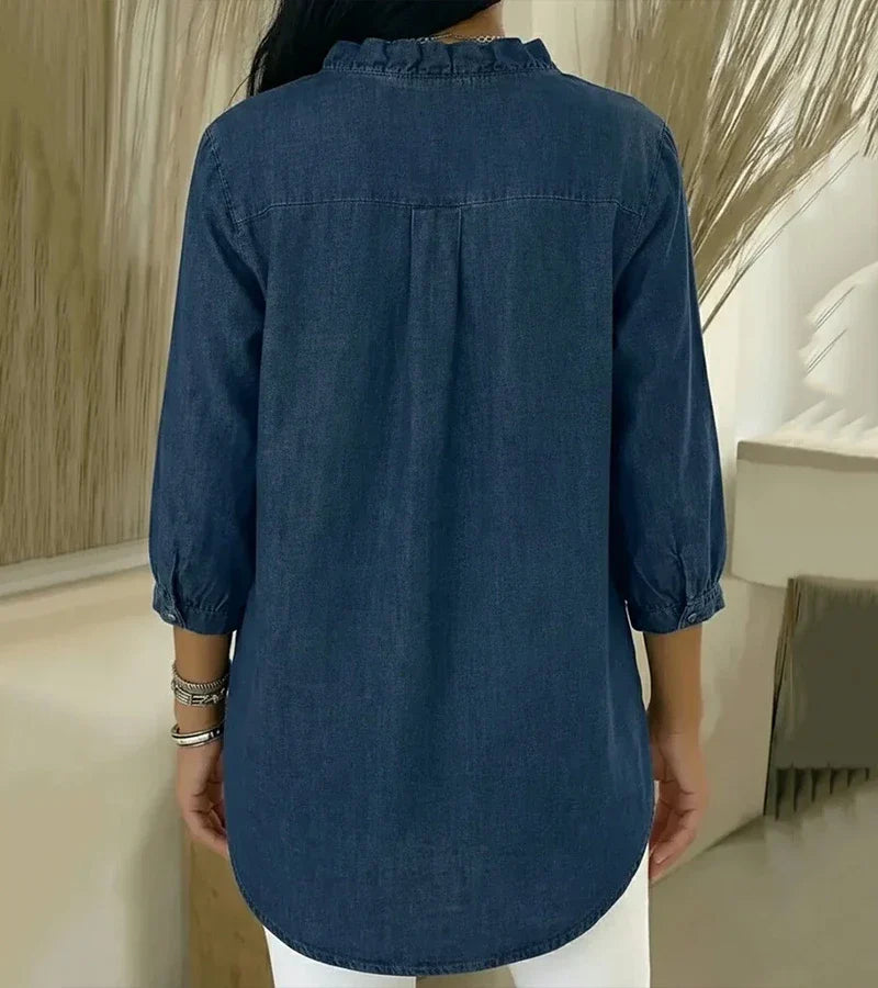 Back view of denim blouse showing ruffled collar and relaxed fit, set against a neutral indoor background.