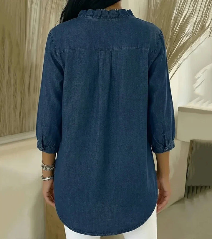 Back view of denim blouse showing ruffled collar and relaxed fit, set against a neutral indoor background.