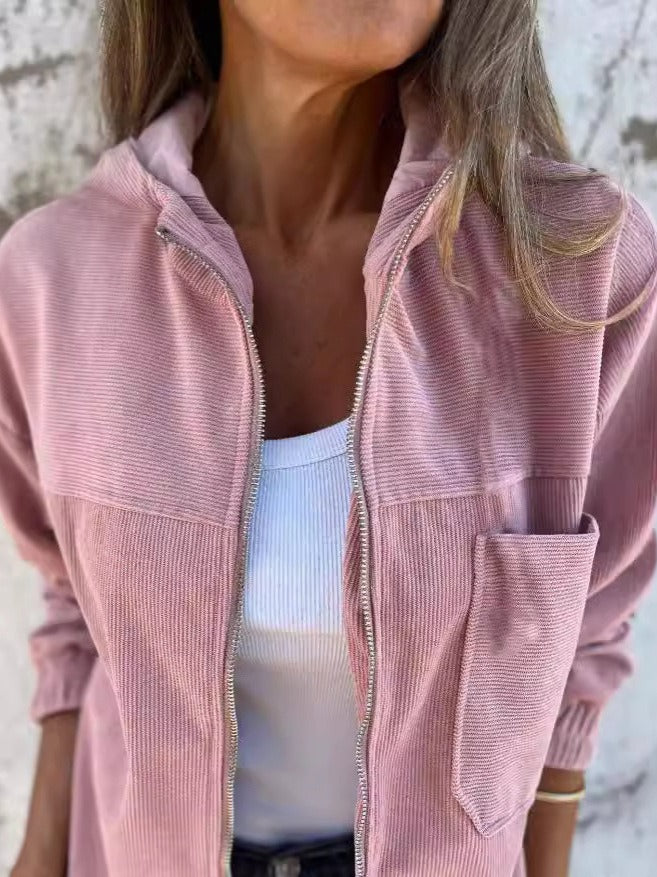 Pink corduroy zip jacket with a white top, highlighting a relaxed silhouette and pockets, set against a rough wall texture.