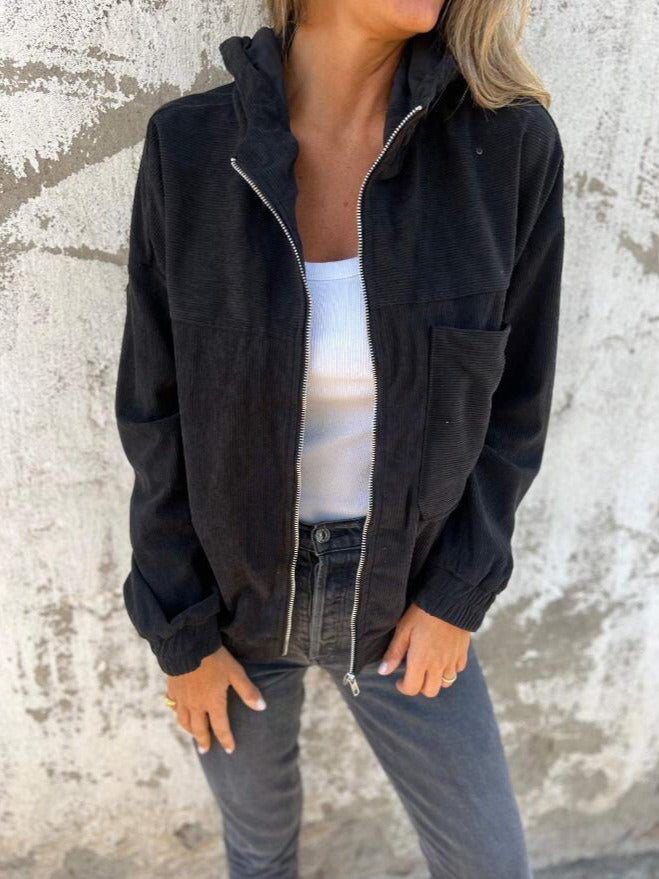 Black corduroy zip jacket worn with a white top, displaying a casual fit and pocket design, in front of a textured wall.