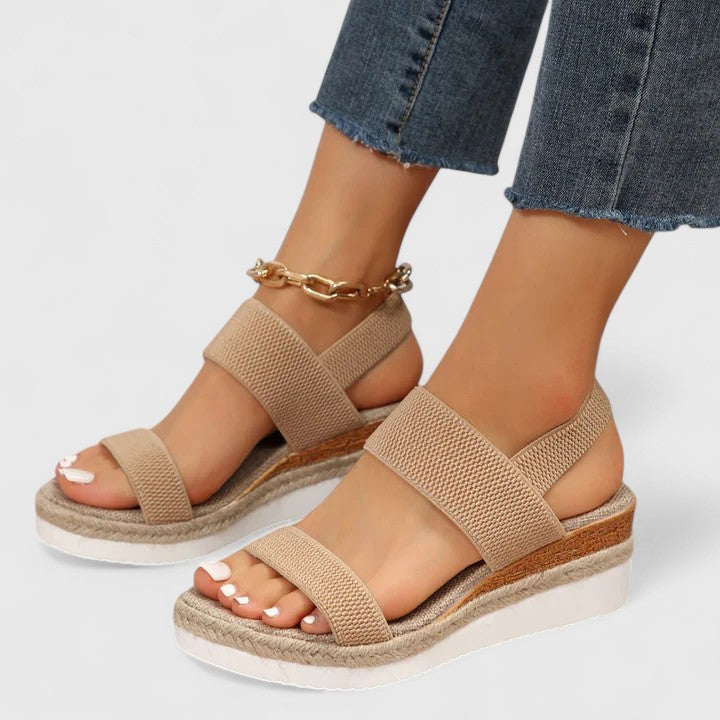 Beige orthopedic sandals with double straps and layered soles, paired with blue jeans and a gold anklet in a studio setting.