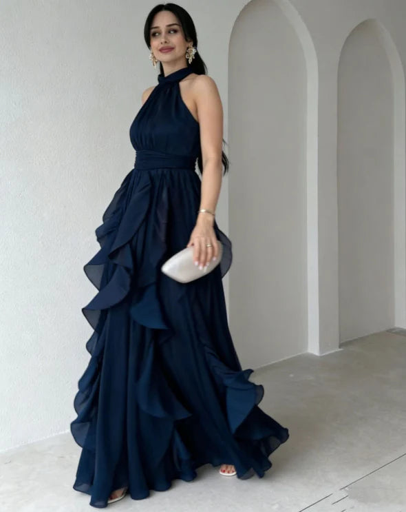 Model wearing a dark navy high neck maxi dress with gathered ruffle layers, holding a clutch, indoors against a white wall.