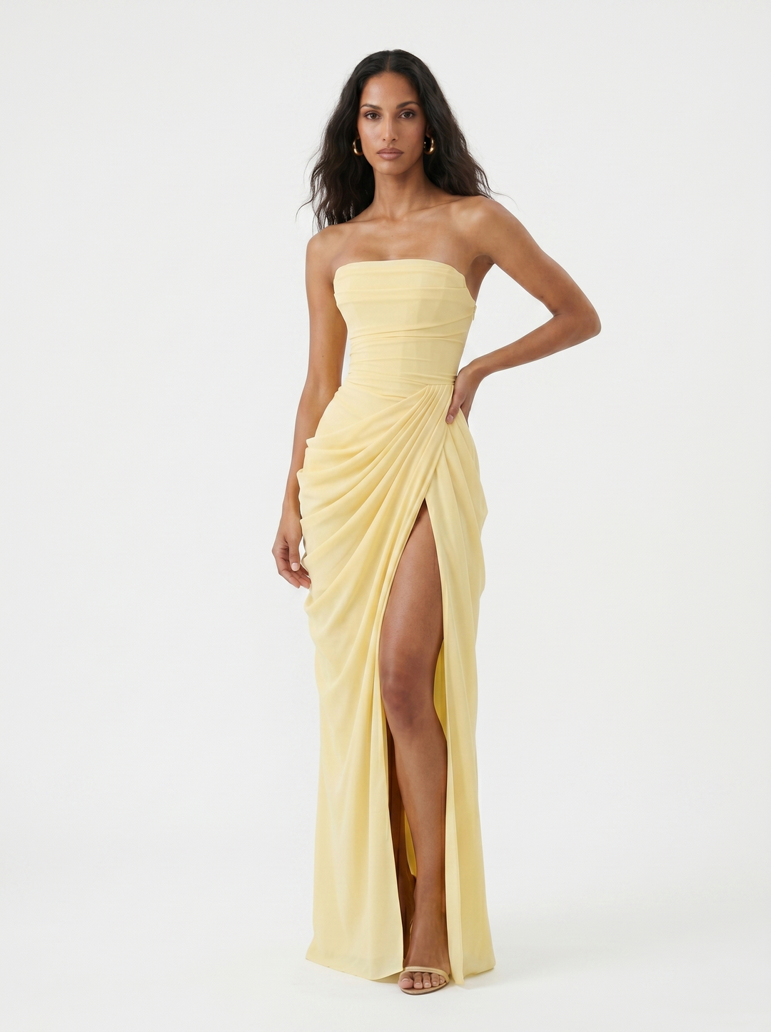 Model posing in a light yellow strapless dress with ruching and a high slit, on a plain studio background.