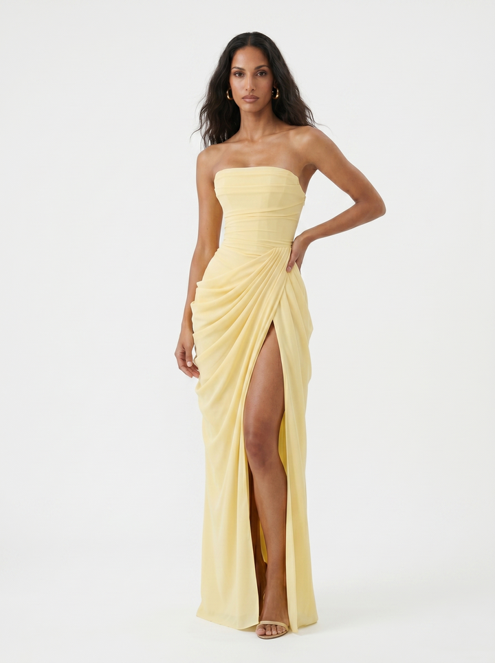 Model posing in a light yellow strapless dress with ruching and a high slit, on a plain studio background.
