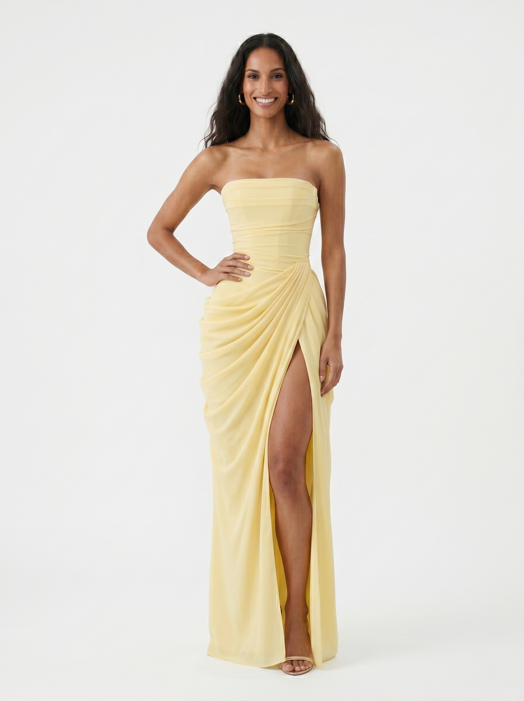 Model smiling in a light yellow strapless dress with ruched details and a high slit, in a studio setting.