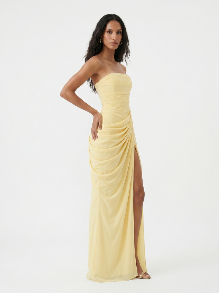 Side view of a model in a light yellow strapless dress with ruching and a high slit, set in studio.