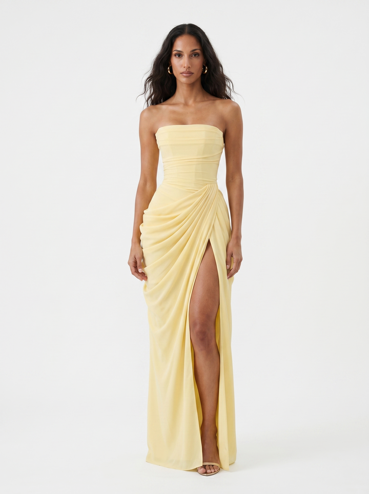 Model wearing a light yellow strapless dress with a ruched design and thigh-high slit in a studio setting.