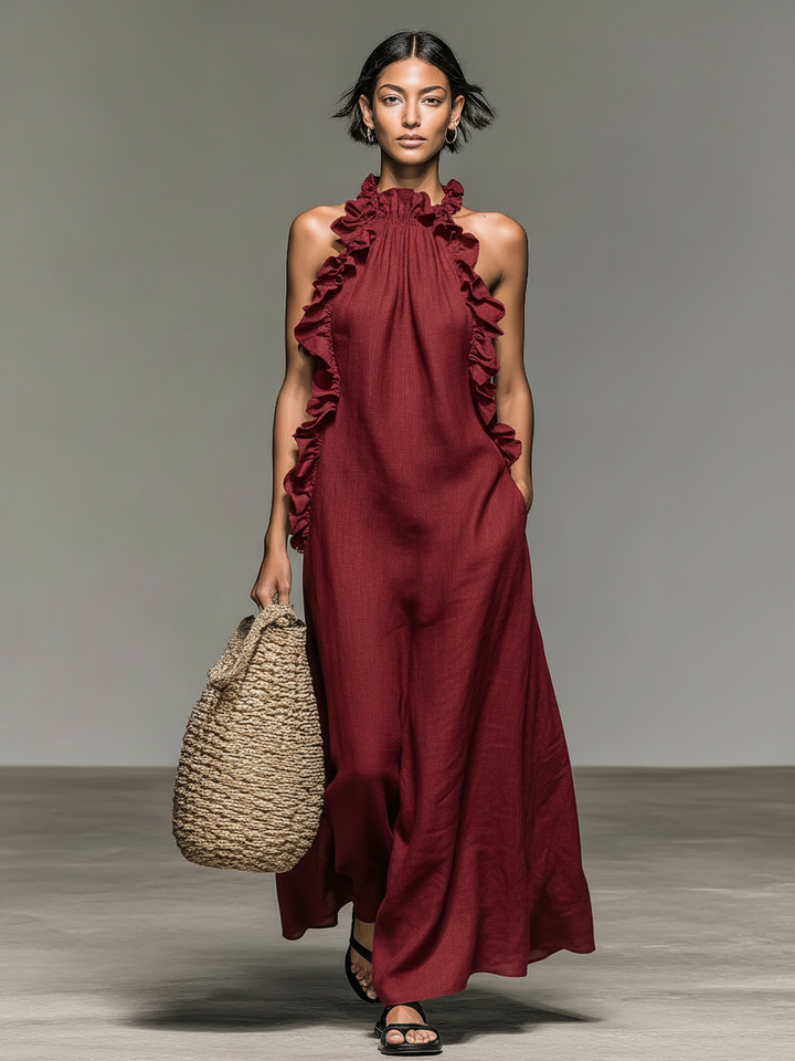 Model in burgundy maxi dress with ruffled details, holding a woven bag, on a concrete floor with gray backdrop.