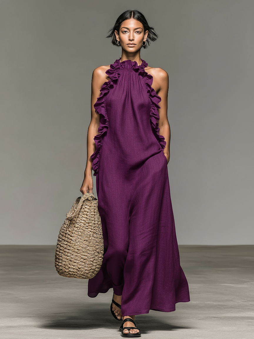 Model in purple maxi dress with ruffles, holding a woven bag, standing on a concrete floor with a gray background.