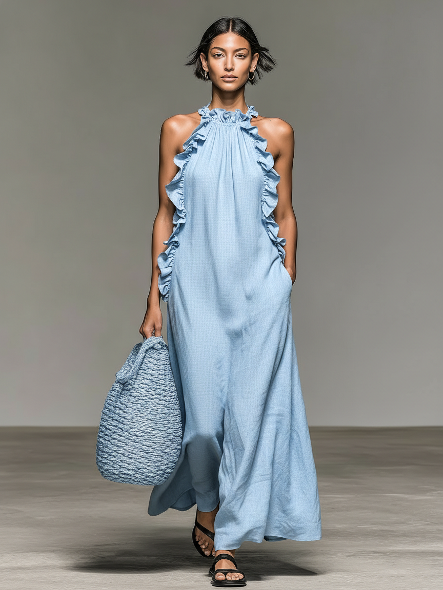 Model wearing a light blue maxi dress with ruffles, holding a matching woven bag, standing on concrete with a gray backdrop.