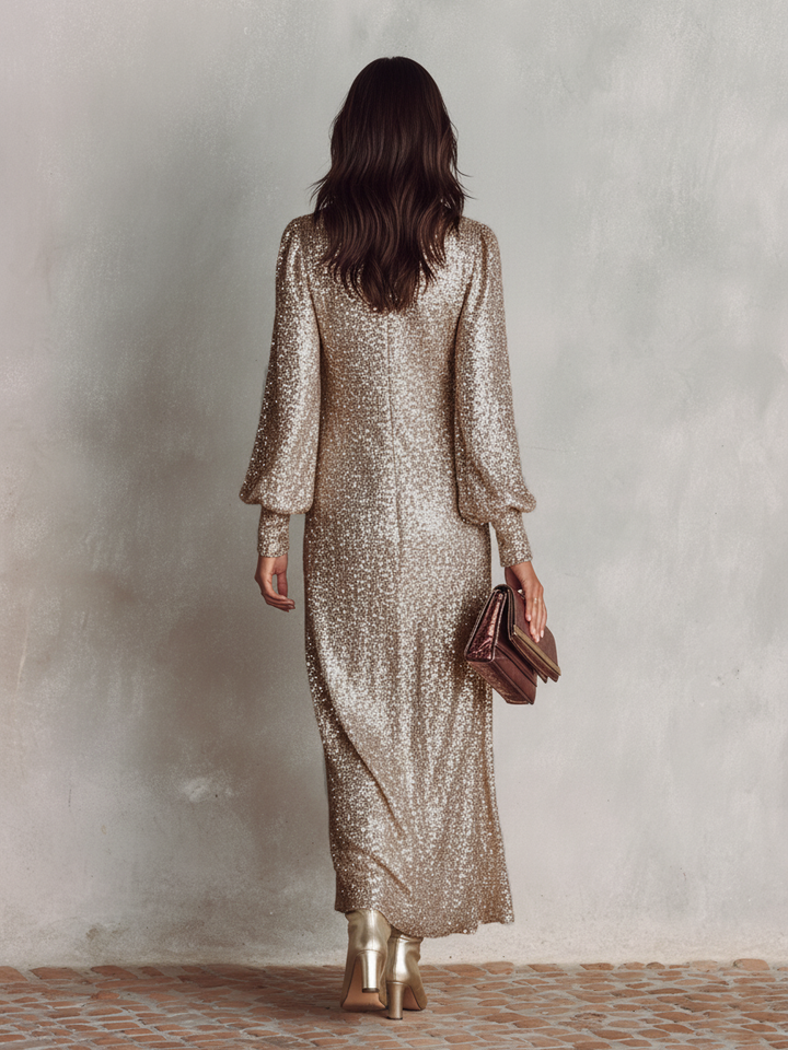 Back view of a woman in a shimmering silver maxi dress with long sleeves, holding a clutch, standing on a stone floor.