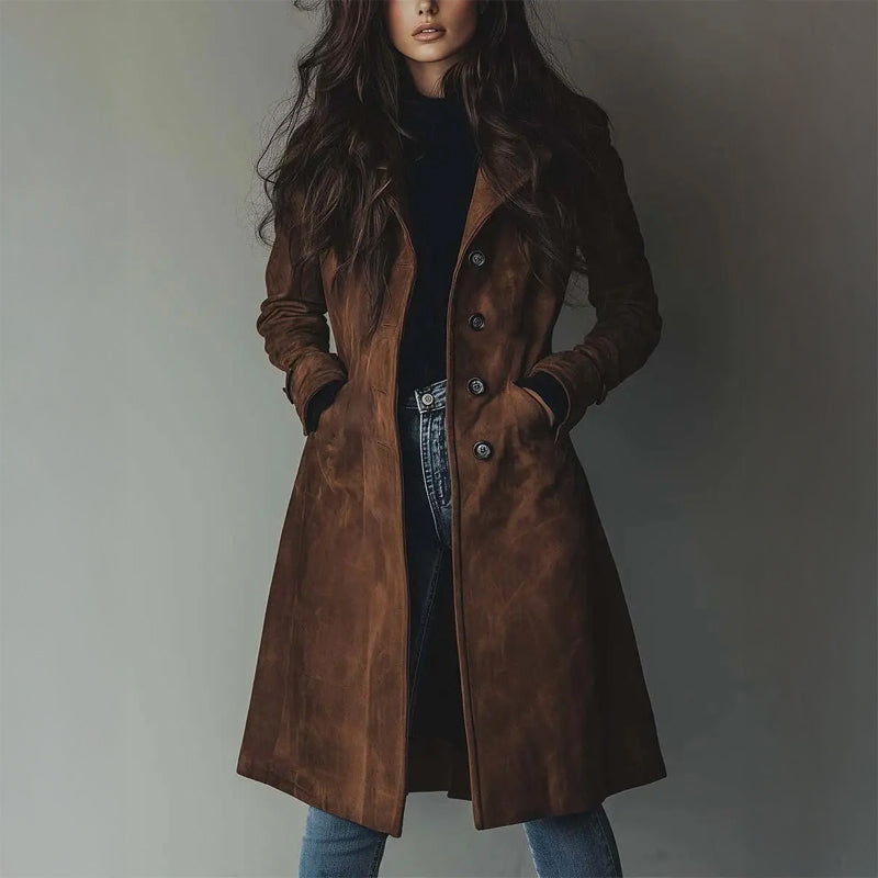 Woman wearing a long, brown coat with buttons, standing against a gray studio background. Hands in pockets.