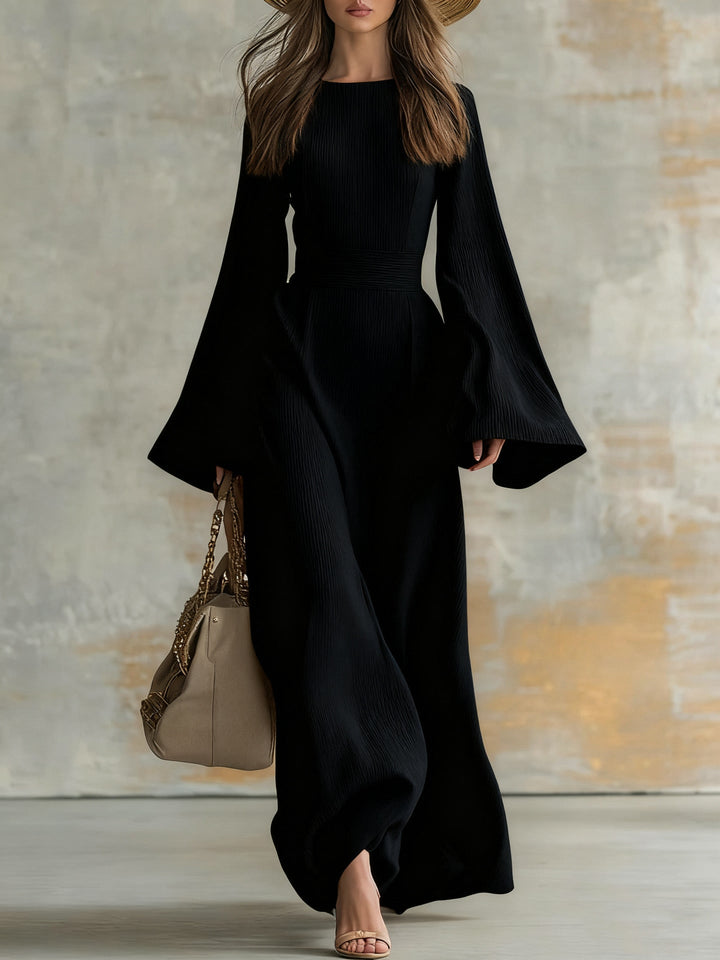 Elegant woman in a long, flowing black dress with flared sleeves, carrying a beige handbag in a studio setting.