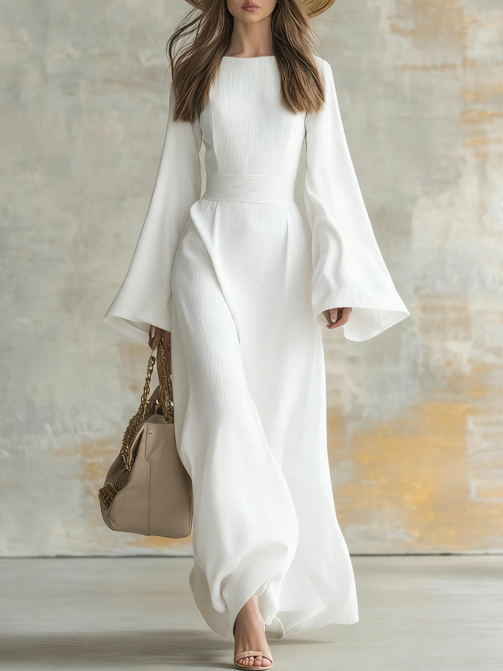 Elegant woman in a long, flowing white dress with flared sleeves, carrying a beige handbag in a studio setting.