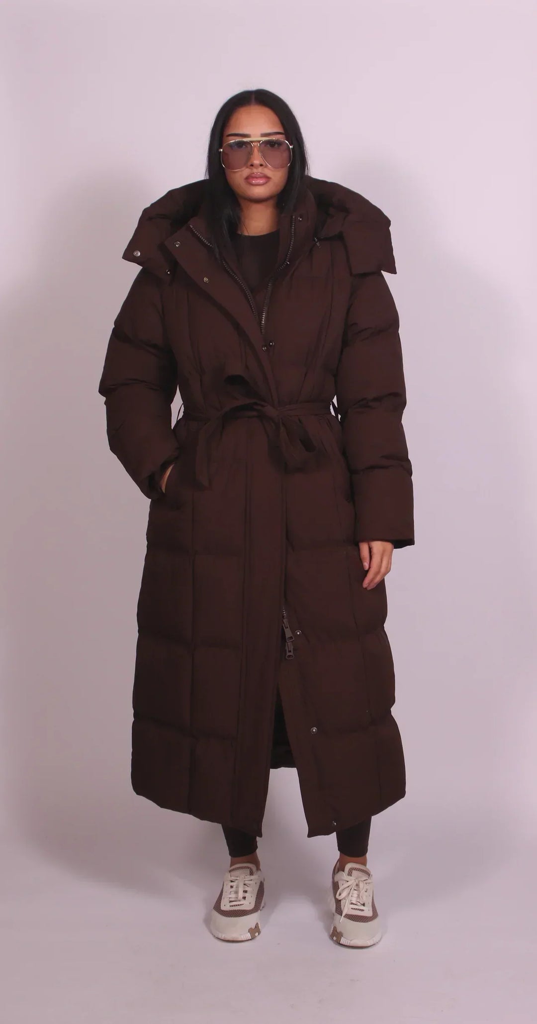 Model wearing a dark brown puffer coat with a belt and hood, standing in a studio with a neutral background.