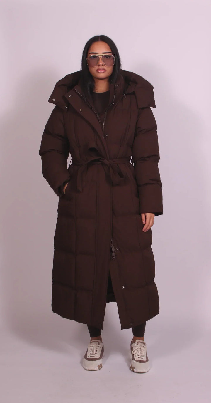 Model wearing a dark brown puffer coat with a belt and hood, standing in a studio with a neutral background.