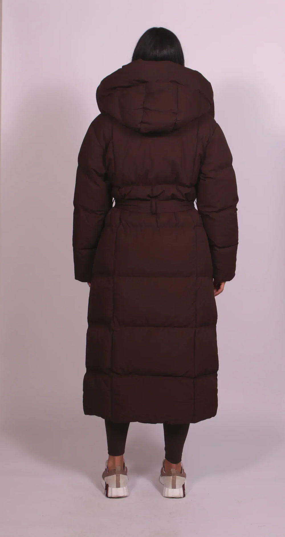 Back view of a model wearing a dark brown puffer coat with a hood and belt, in a simple studio setting.