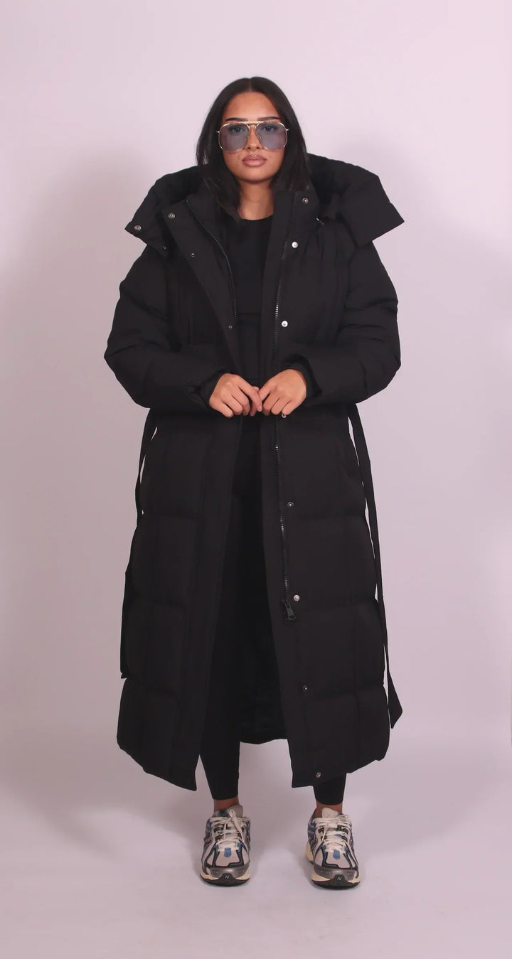 Model in a black puffer coat with buttons, hood, and zipper, standing in a studio with a plain backdrop.