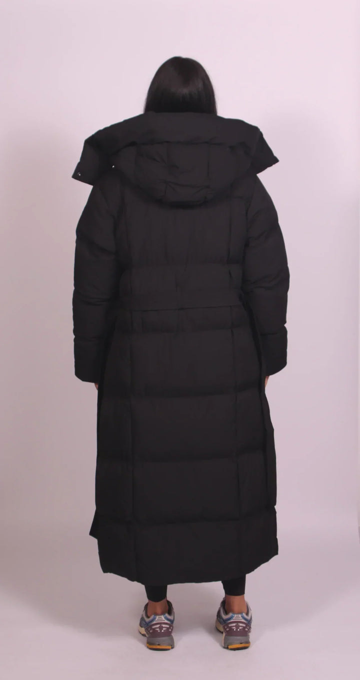 Back view of a black puffer coat with hood and belt, worn by a model in a minimalistic studio environment.